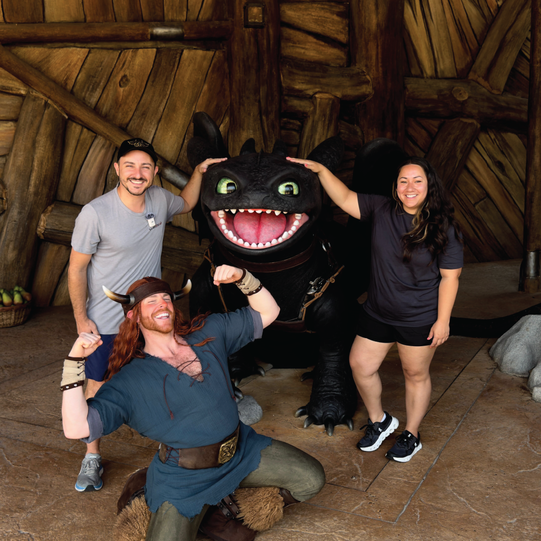 Three people posing with a large, smiling black dragon character inside a wooden-themed setting; one person is dressed in Viking-style costume kneeling and flexing muscles.