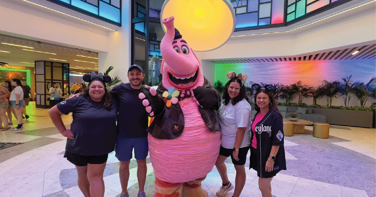 Four people wearing Mickey and Minnie Mouse ears standing indoors around a large pink elephant character in a colorful lobby.