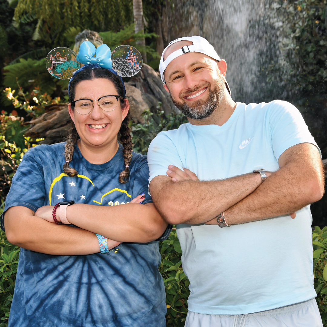 Smiling man and woman standing side by side with arms crossed in a garden setting, with the woman wearing Mickey Mouse ears.