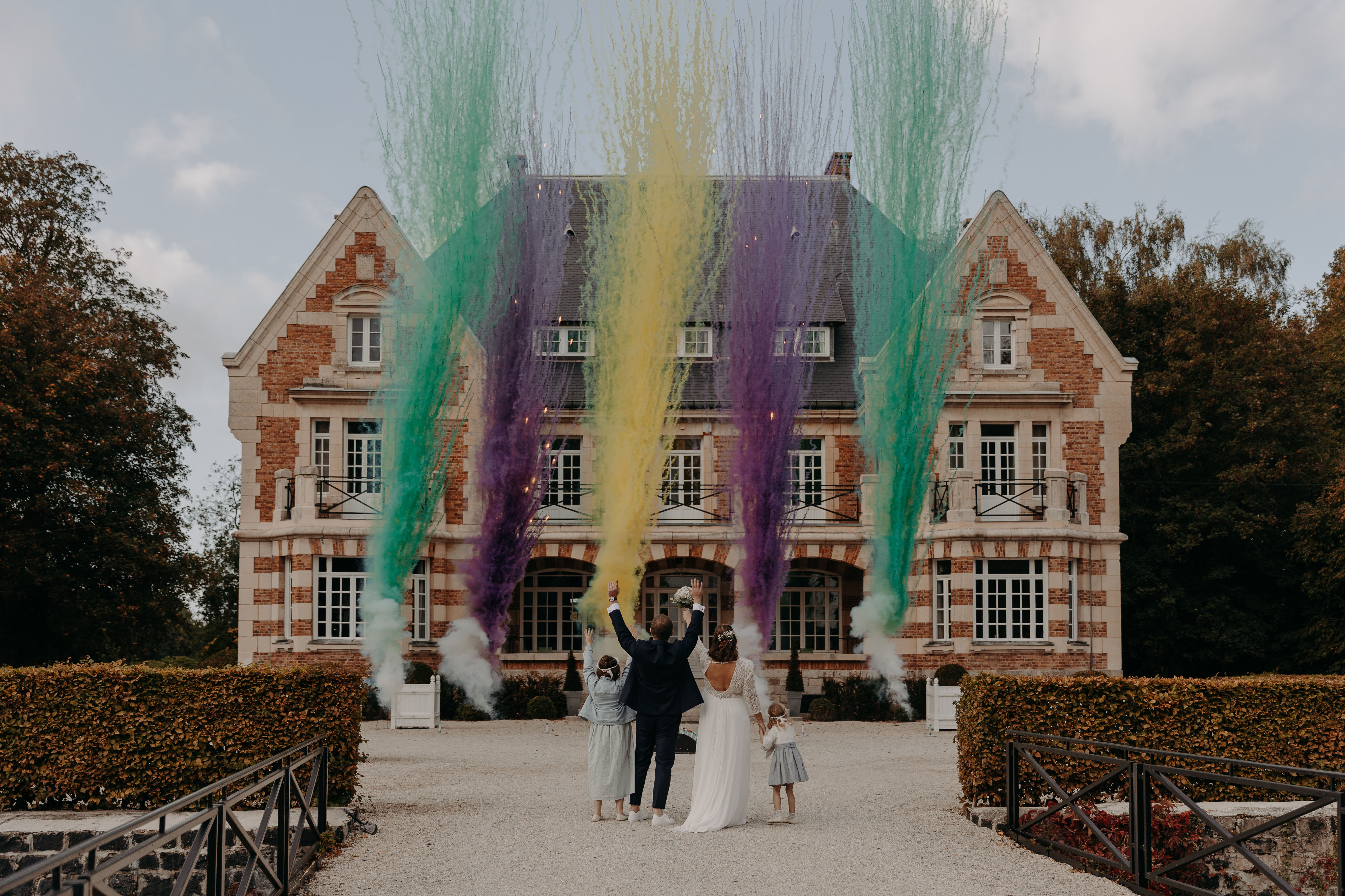 Famille de mariés bras levés devant un château avec jets de fumées vertes jaunes et violettes, artifices de jour mariage Wagnon