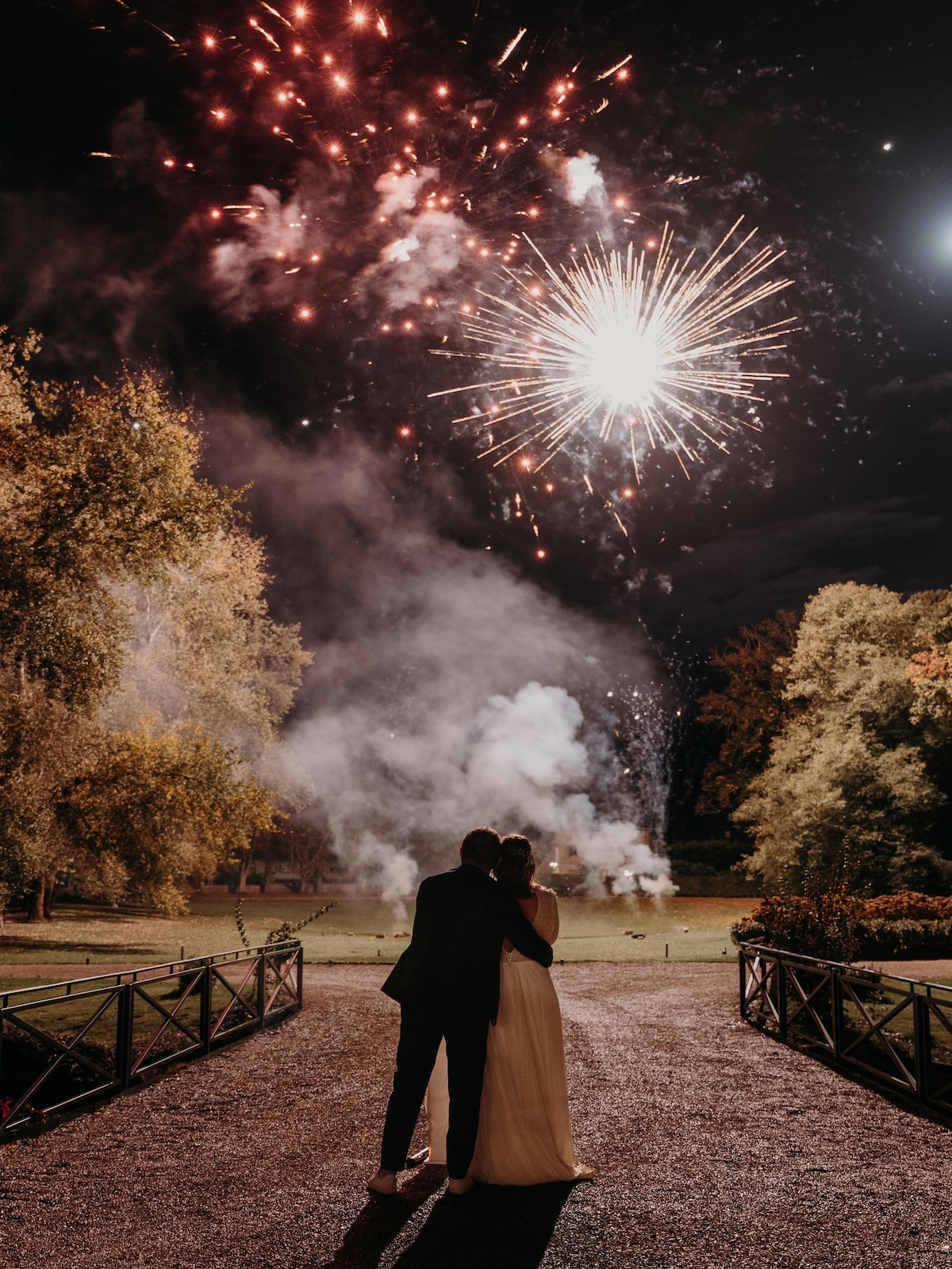 Couple de mariés qui regardent le feu d'artifice coloré explosant dans le ciel nocturne, spectacle pyrotechnique mariage Wagnon Events