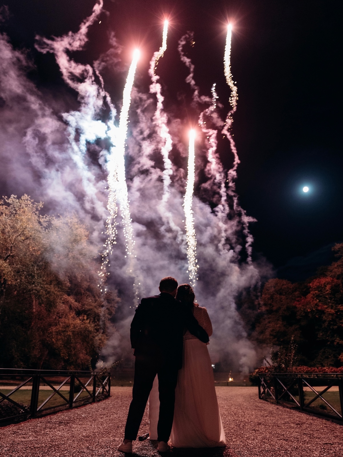 Couple de mariés regardant des fusées lumineuses blanches et roses s'élever dans le ciel nocturne, feux d'artifice mariage Wagnon Events
