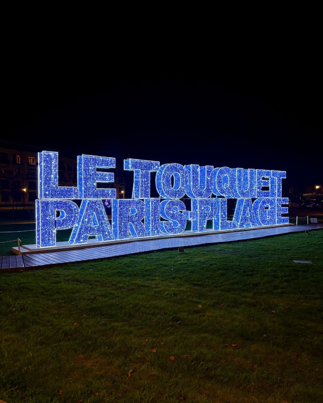 Installation géante "Le Touquet Paris-Plage" réalisée en lettres illuminées de LED bleues, photographiée de nuit sur la pelouse du front de mer.