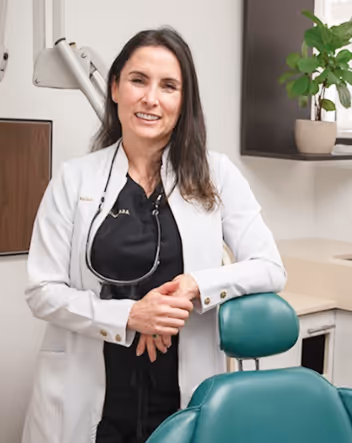 Female dentist in white coat standing and smiling beside a dental chair in a clinic room.
