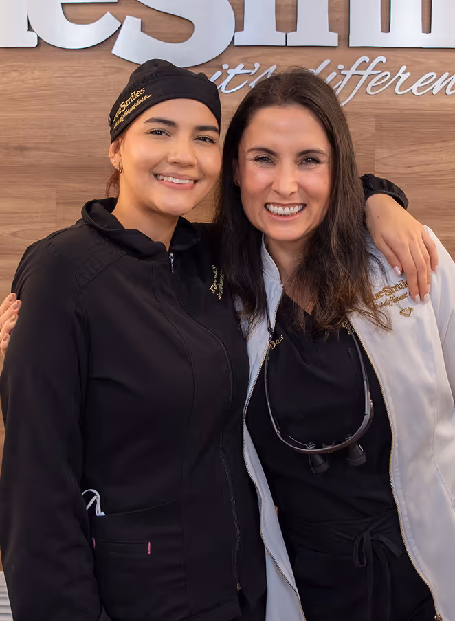 Two female dental professionals smiling and posing together, one in black scrubs and the other in a white jacket with dental magnifying glasses around her neck.