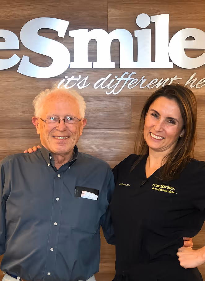 An elderly man with glasses and white hair smiles next to a woman in black scrubs with 'truesmiles' embroidered, standing in front of a wooden wall with large 'Smile' text.