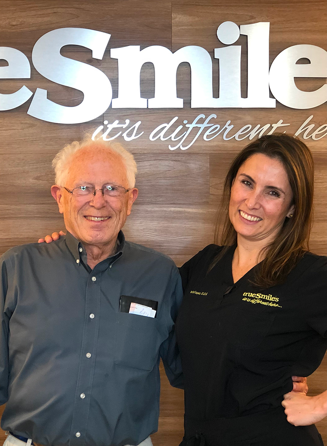 An elderly man with glasses and white hair smiles next to a woman in black scrubs with 'truesmiles' embroidered, standing in front of a wooden wall with large 'Smile' text.