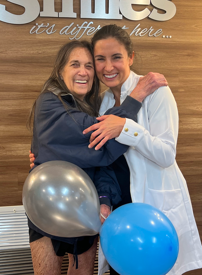 Two women smiling and hugging, one holding a silver balloon and the other a blue balloon, standing in front of a wooden wall with text.