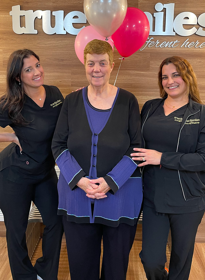Three smiling women standing together in front of a wooden wall with 'TrueSmiles' signage and colorful balloons above.
