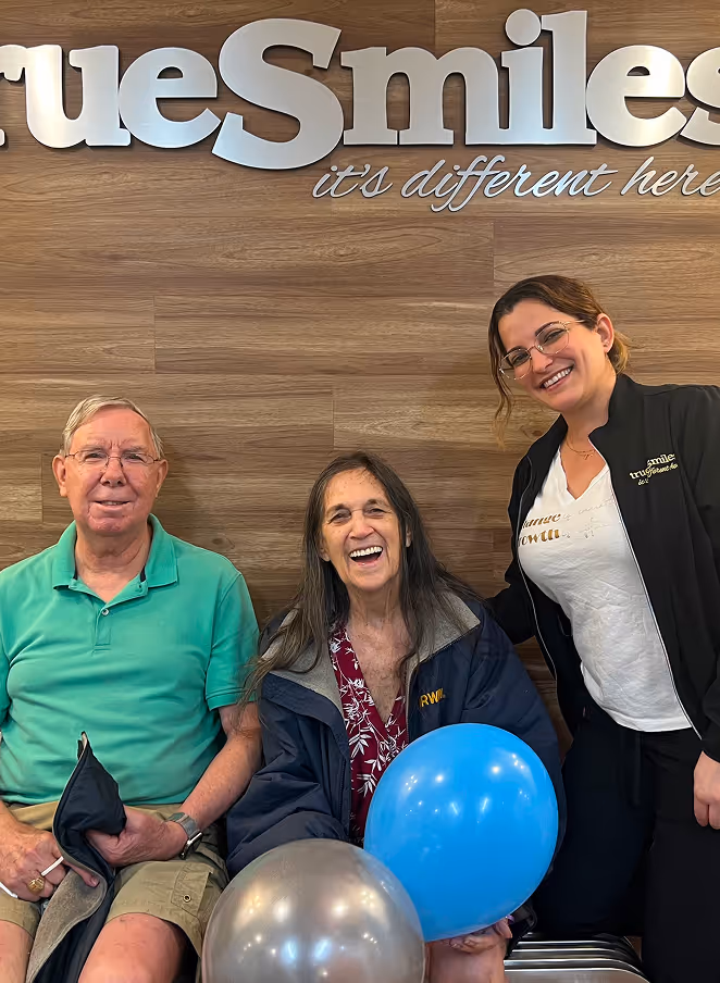 Three people smiling in front of a wooden wall with the silver sign 'TrueSmiles it's different here'; the woman in the middle holds blue and silver balloons.