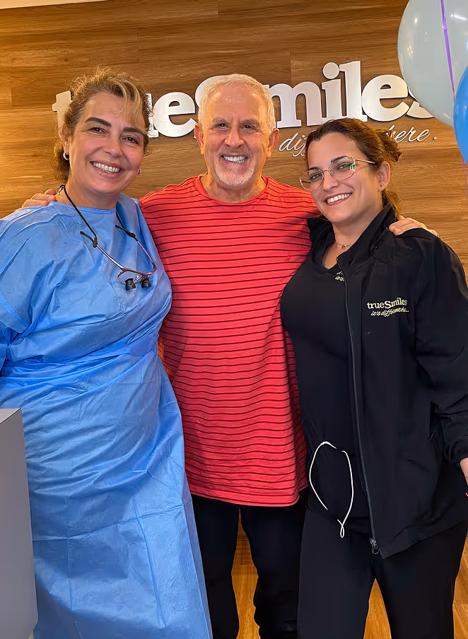 Smiling elderly man in red striped shirt stands between two smiling women, one in blue dental scrubs and the other in black TrueSmiles jacket, indoors with wooden wall background.