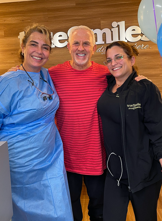 Smiling elderly man in red striped shirt stands between two smiling women, one in blue dental scrubs and the other in black TrueSmiles jacket, indoors with wooden wall background.