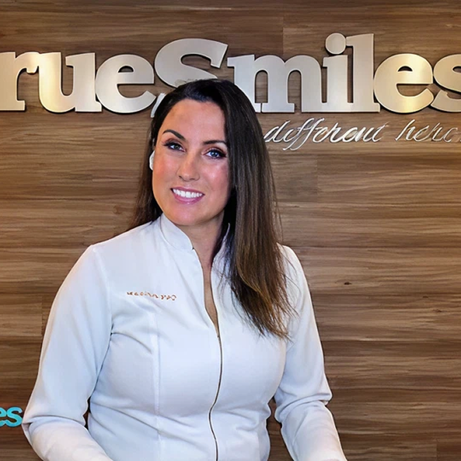 Woman with long brown hair wearing a white zip-up jacket smiling in front of a wooden wall with the sign 'True Smiles'.
