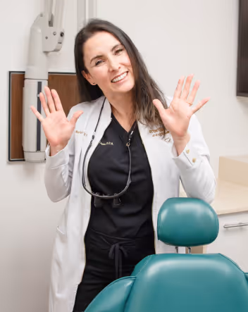 Smiling female dentist in a white coat raising both hands in a dental office.