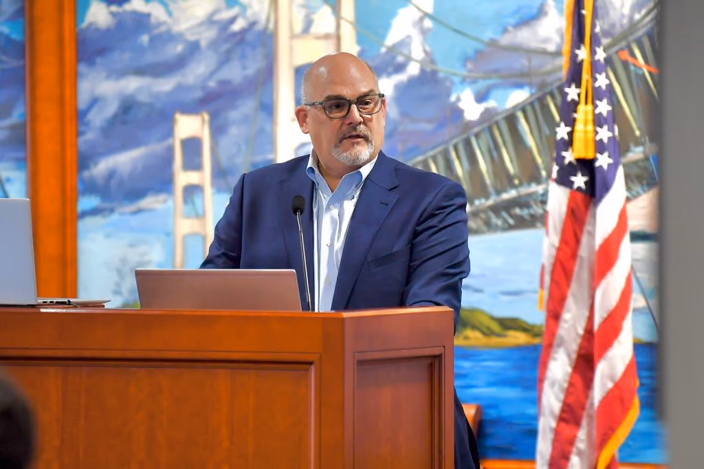 Man in glasses and navy suit speaking at a podium with an American flag and a mural of the Golden Gate Bridge in the background.