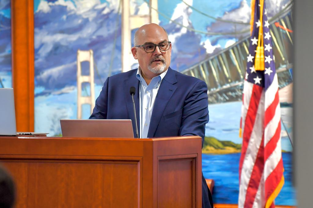 Man in glasses and navy suit speaking at a podium with an American flag and a mural of the Golden Gate Bridge in the background.