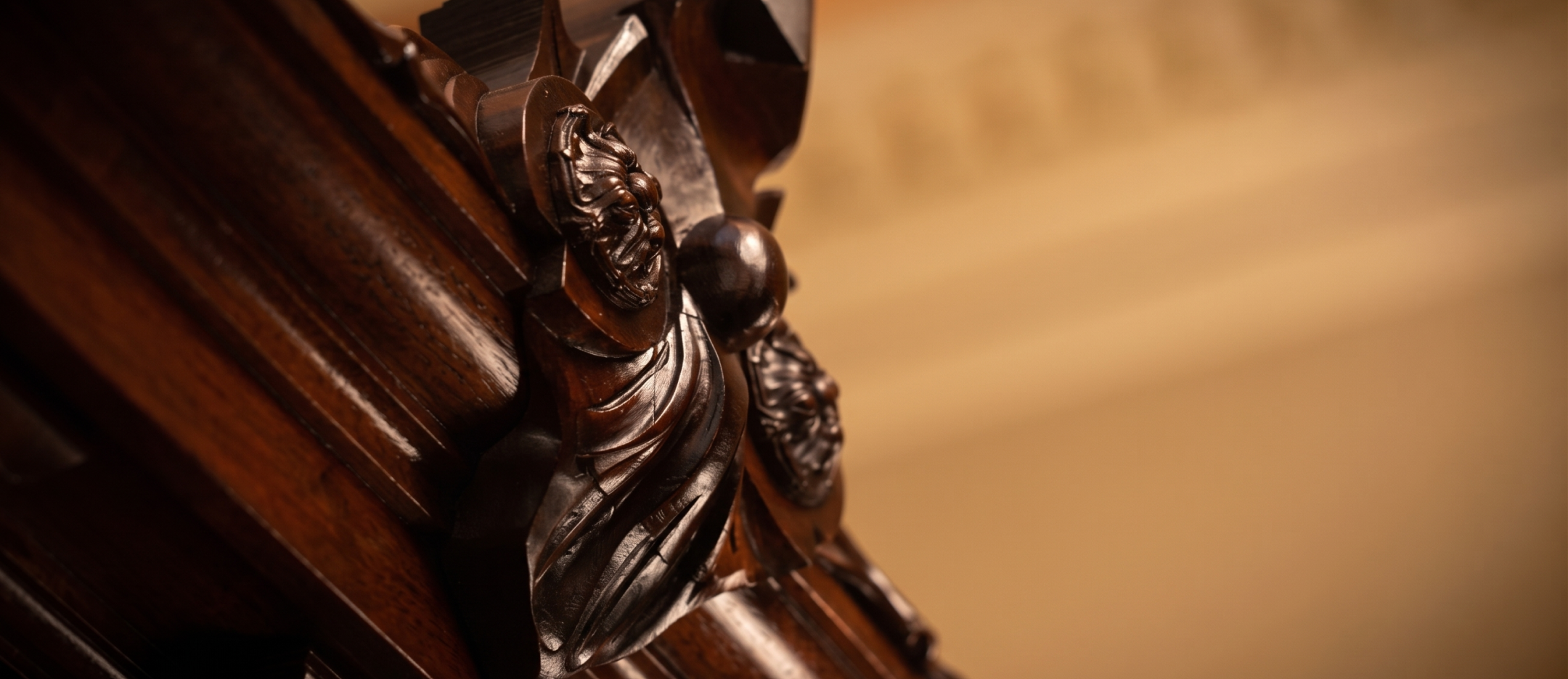 Close-up of detailed dark wood carving with floral and flowing patterns on a polished wooden surface, against a blurred beige background.