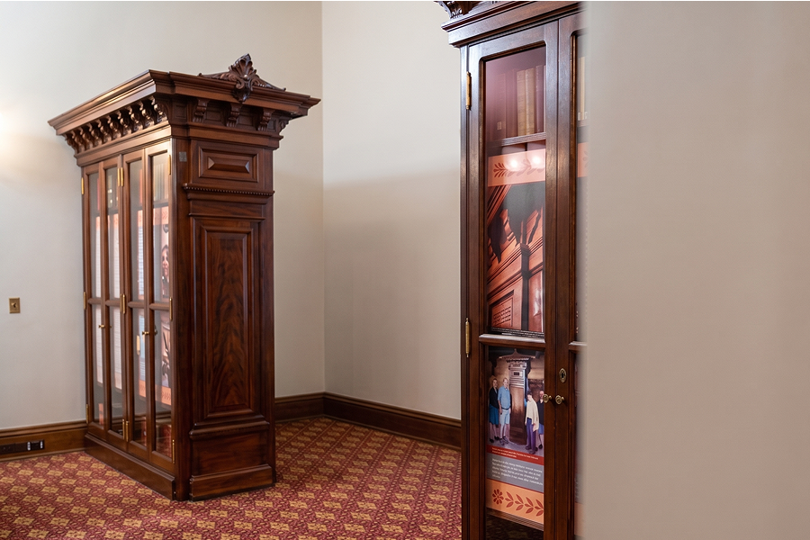 Two ornate wooden display cabinets with glass doors standing on a patterned red carpeted floor inside a room with beige walls. The cabinet glass shows images of people and architectural elements inside.