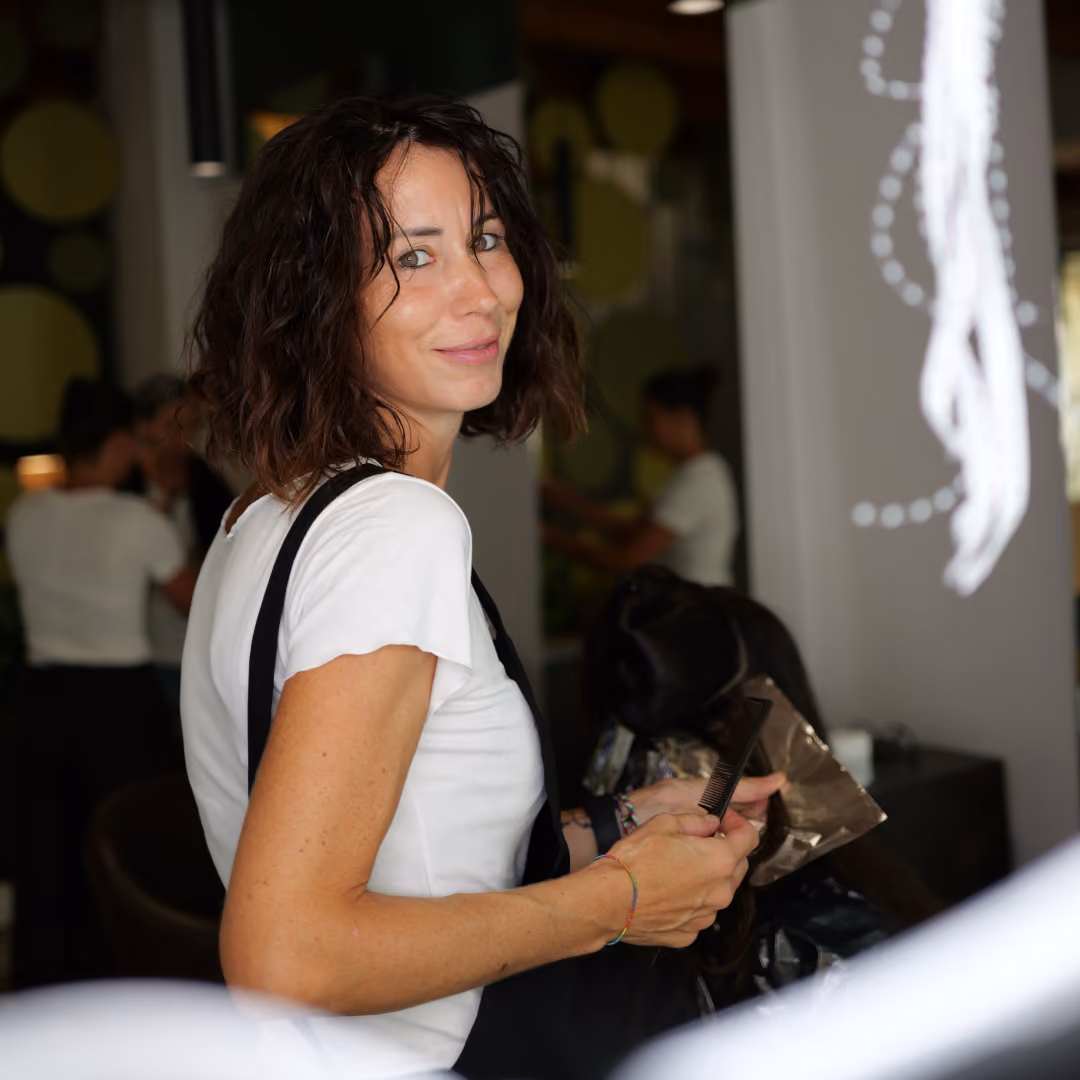 Ritratto in salone di una stilista del salone Gala con capelli scuri mossi, sorridente e pronta per un trattamento.