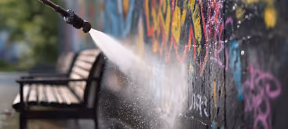 Hochdruckreiniger entfernt bunte Graffiti von einer Wand im Freien nahe einer Bank.
