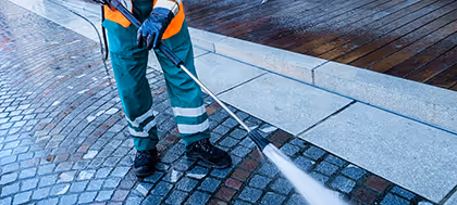Arbeiter in Warnkleidung reinigt nasses Pflaster mit Hochdruckreiniger vor Gehweg.