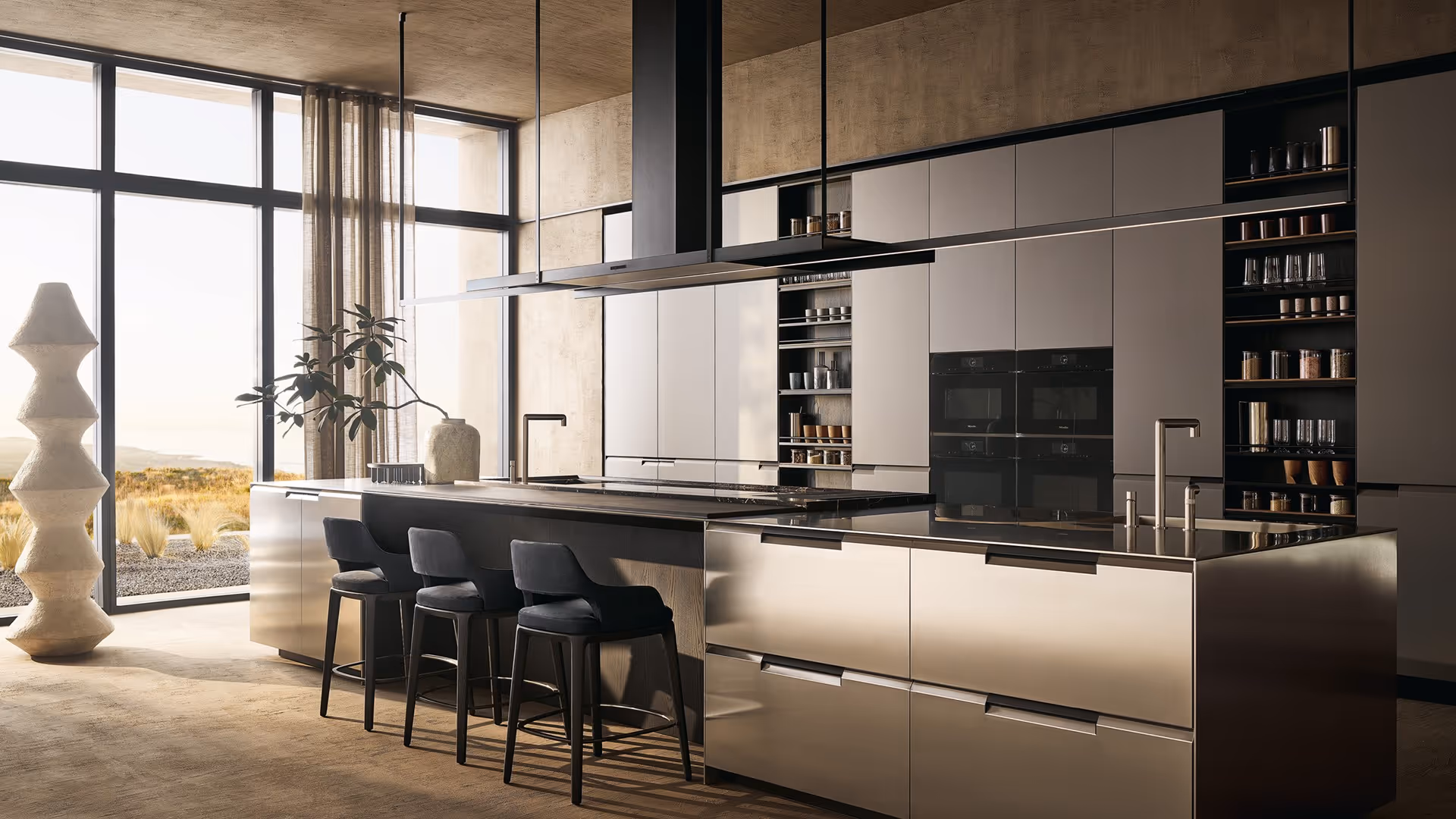 Modern kitchen with large island, three black bar stools, sleek beige cabinets, built-in ovens, open shelving, and floor-to-ceiling windows overlooking a natural landscape.