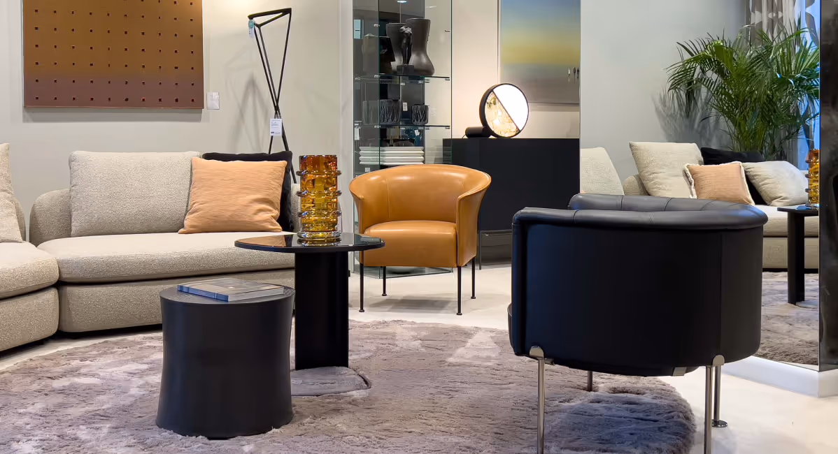 Modern living room with beige sofa, tan leather chair, black armchair, round black side tables, and decorative amber glass vase.