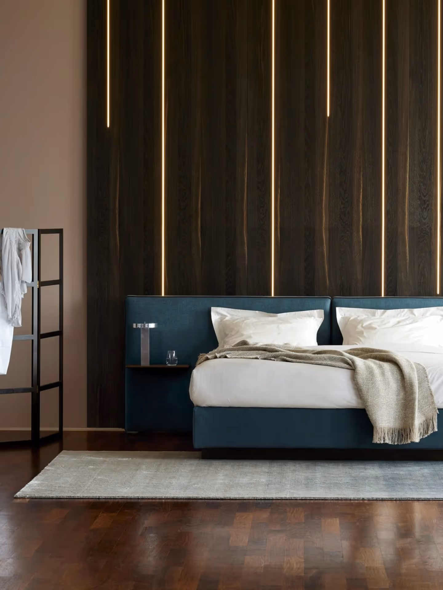 Modern bedroom with a teal upholstered bed, white bedding, beige throw, vertical strip lights on dark wood wall, and a black clothes rack with white clothes.