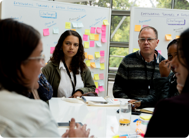 A diverse group of people engaged in a meeting around a table with whiteboards covered in colorful sticky notes behind them.