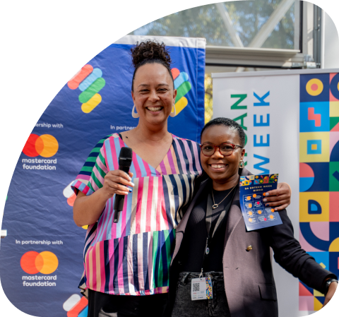 Two smiling women standing together at an event, one holding a microphone and the other holding a colorful card, with Mastercard Foundation and African Week banners in the background.