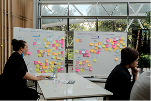 Two people seated at a table in front of whiteboards covered with colorful sticky notes in a bright room with large windows.