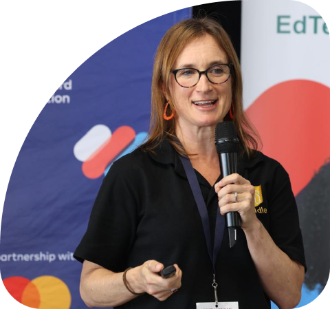 Two smiling women standing together at an event, one holding a microphone and the other holding a colorful card, with Mastercard Foundation and African Week banners in the background.