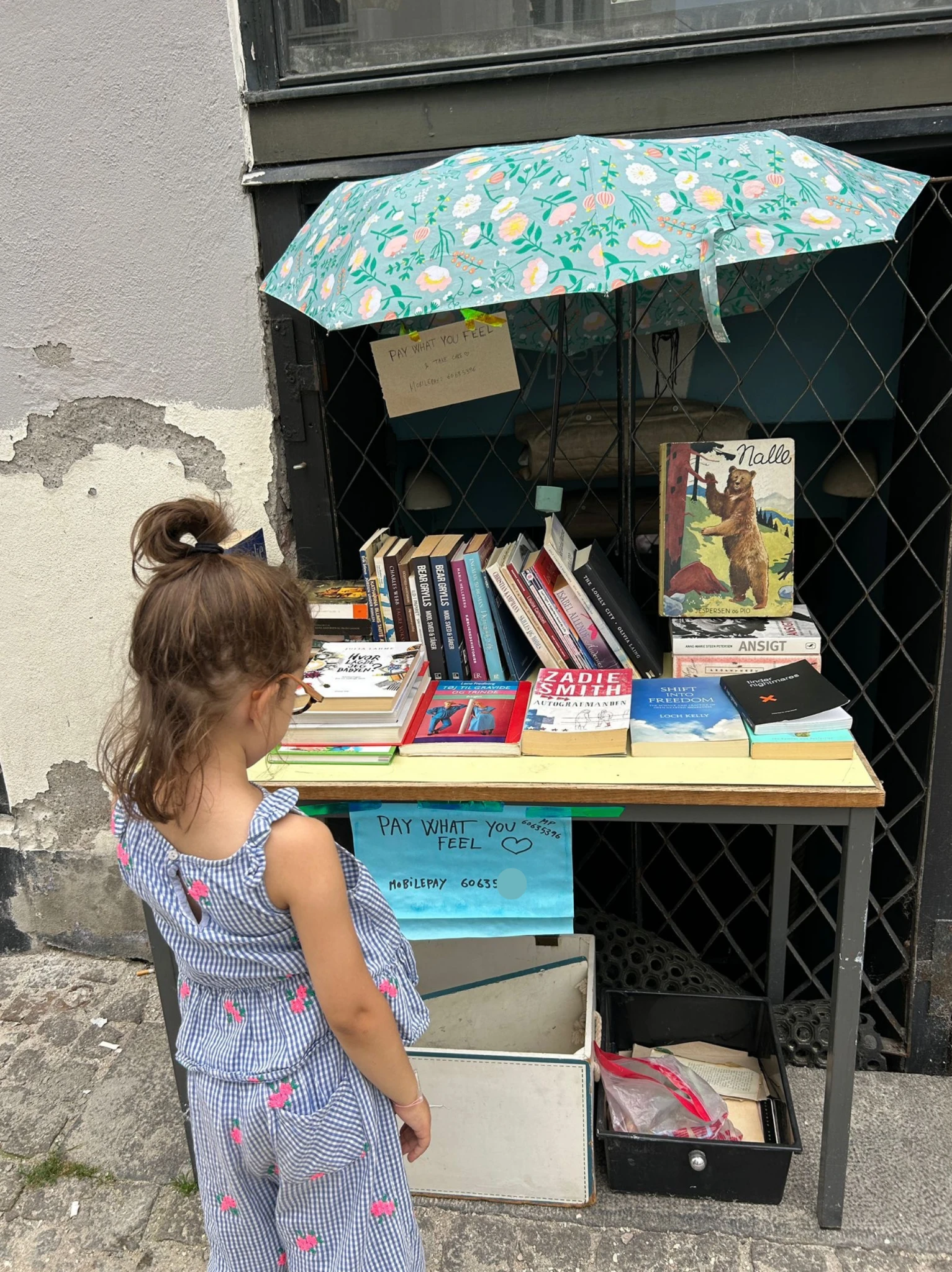 À Copenhague, au Danemark, une fillette observe un stand de livres de seconde main en libre-service, marché de rue façon vide-grenier, symbole de confiance.