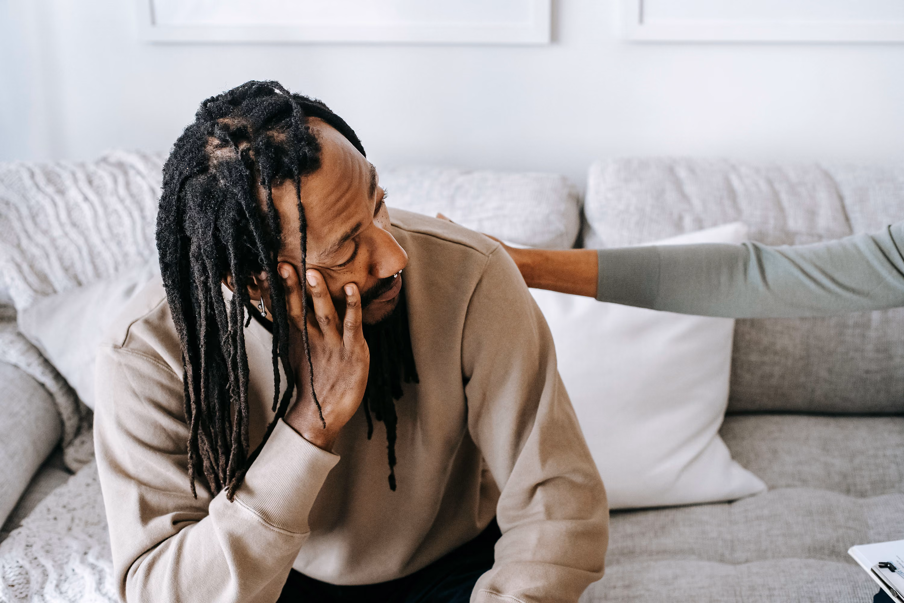 Man sitting on a couch looking down with hand on face while another person consoles him by touching his shoulder.