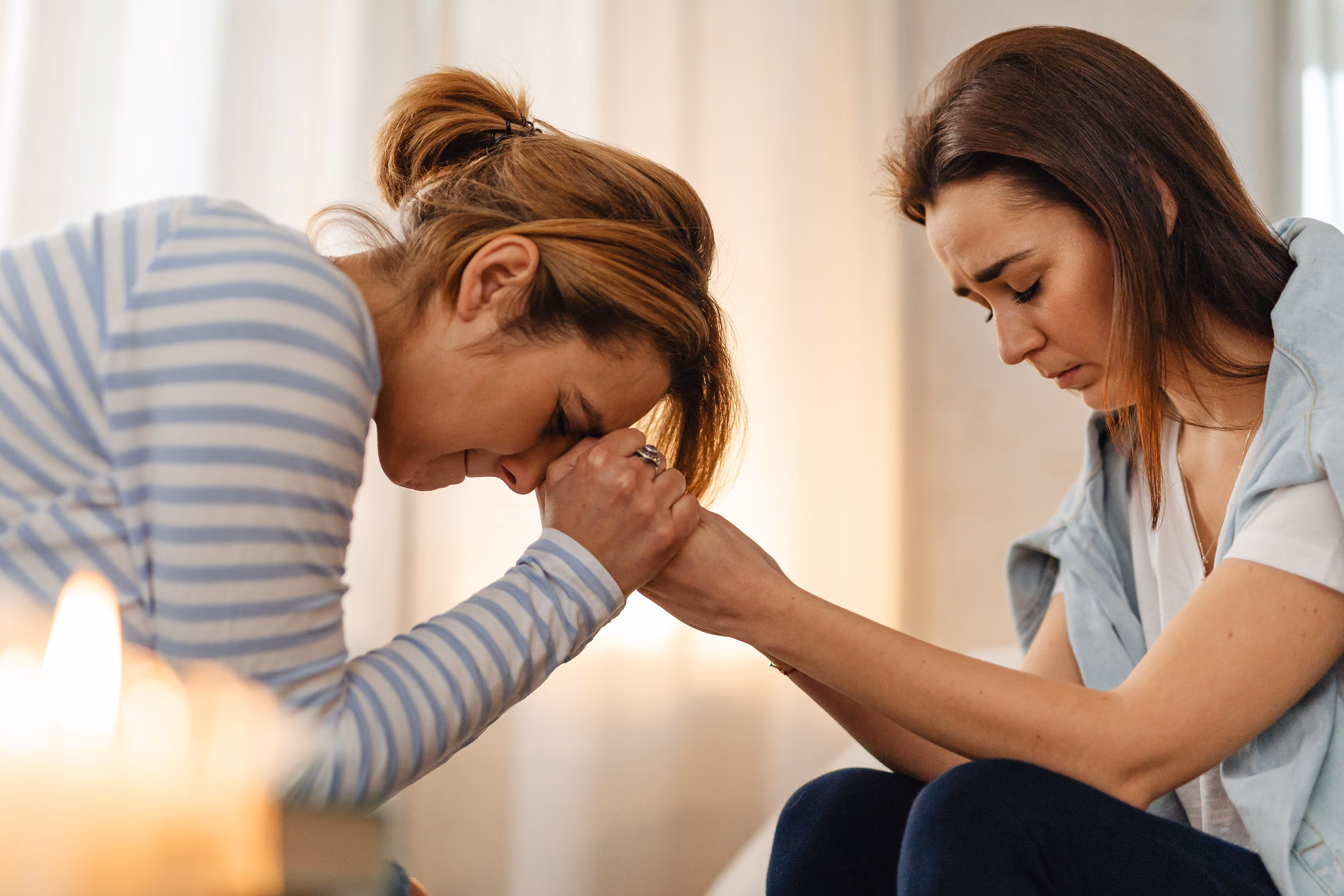 Two women sitting face to face holding hands with eyes closed, consoling each other.