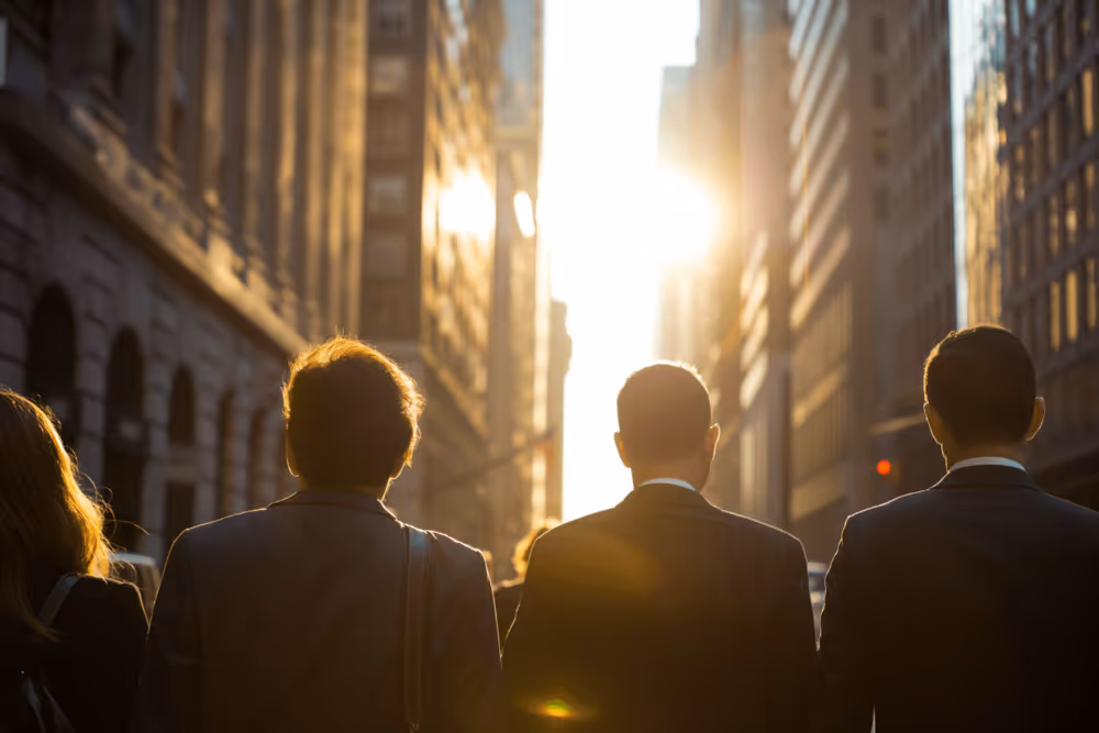 Group of business professionals walking down a city street at sunrise or sunset, surrounded by tall buildings and illuminated by bright sunlight in the distance.
