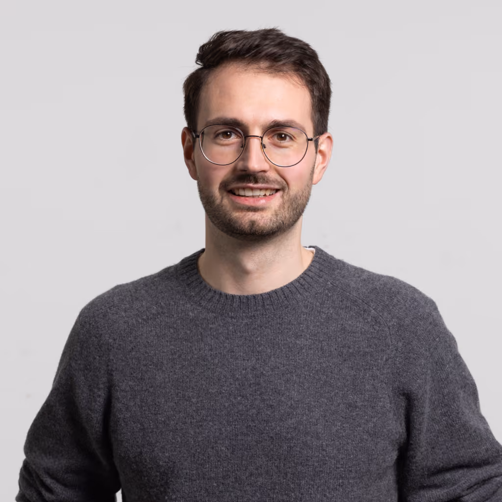 Smiling young man with short brown hair, beard, and round glasses wearing a gray sweater against a plain light background.