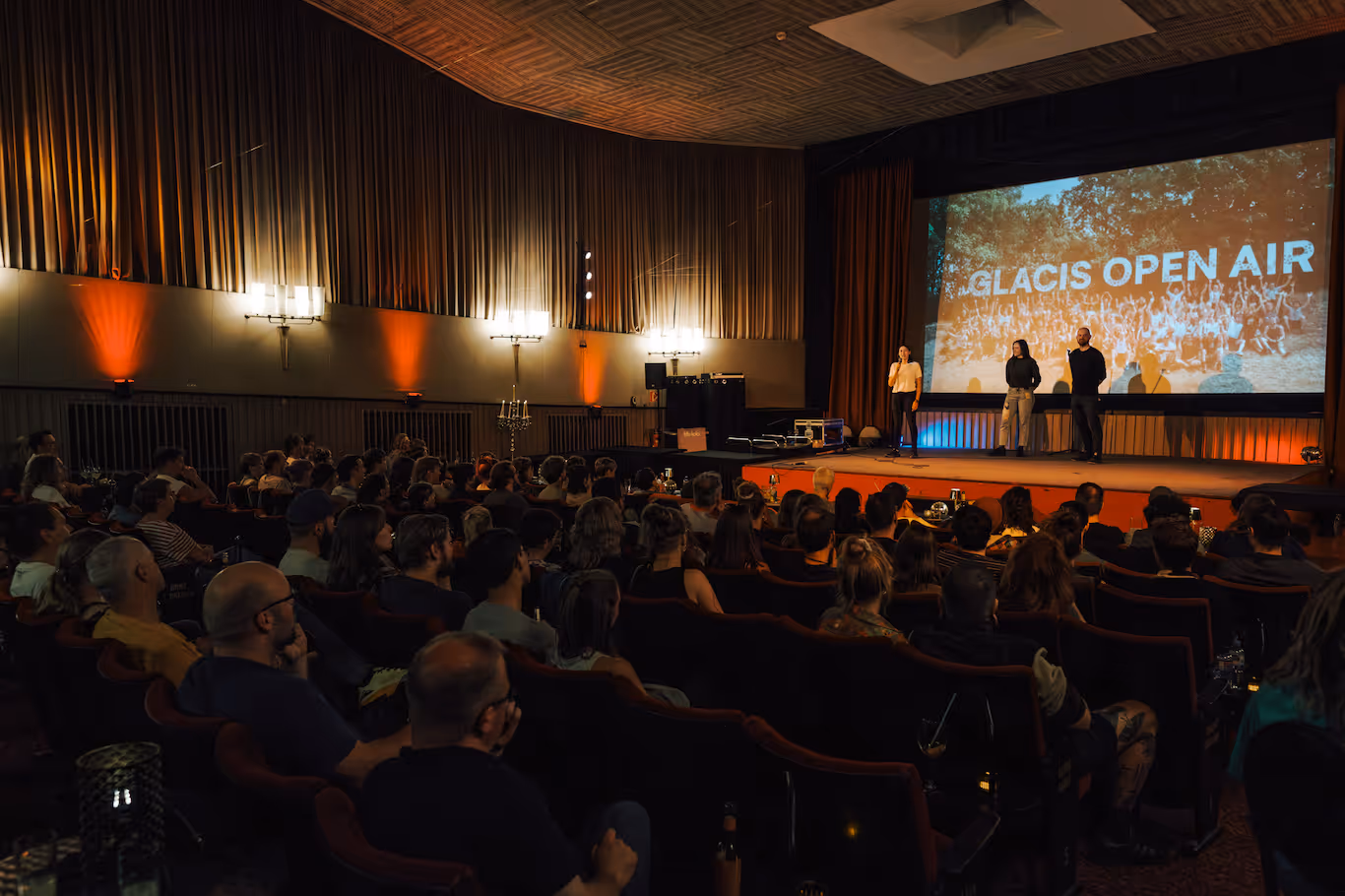 Viele Menschen sitzen in einem Kino, auf der Bühne stehen drei Personen