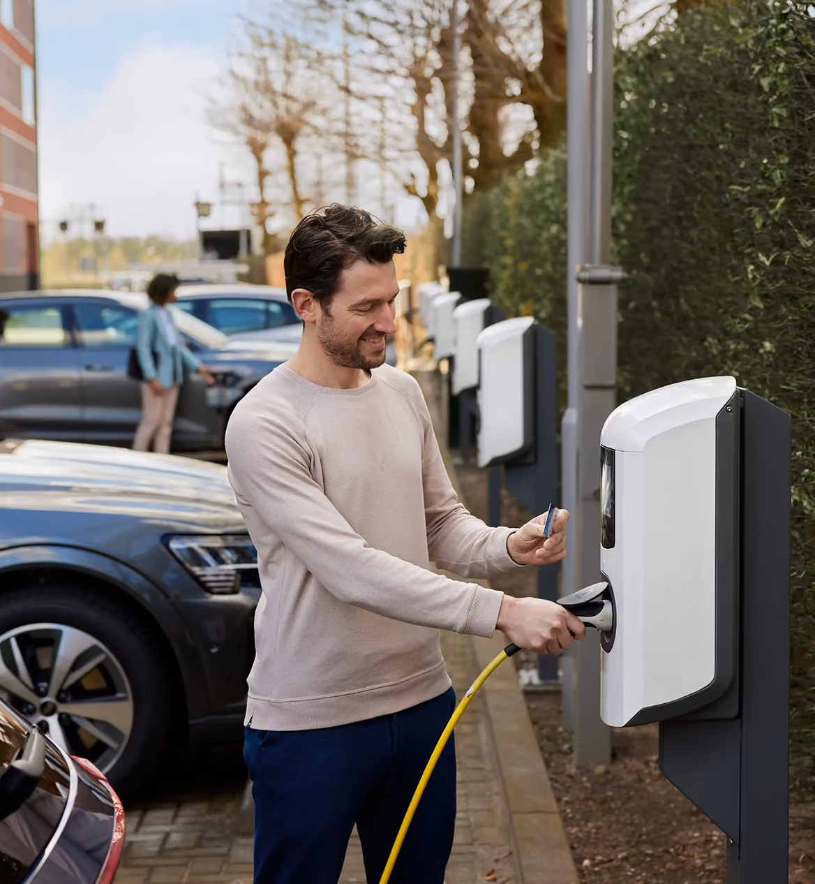Lachende man houdt een laadpas tegen een witte laadpaal terwijl hij de gele laadkabel vasthoudt, met meerdere geparkeerde elektrische auto's en laadpunten op een bedrijfsterrein.