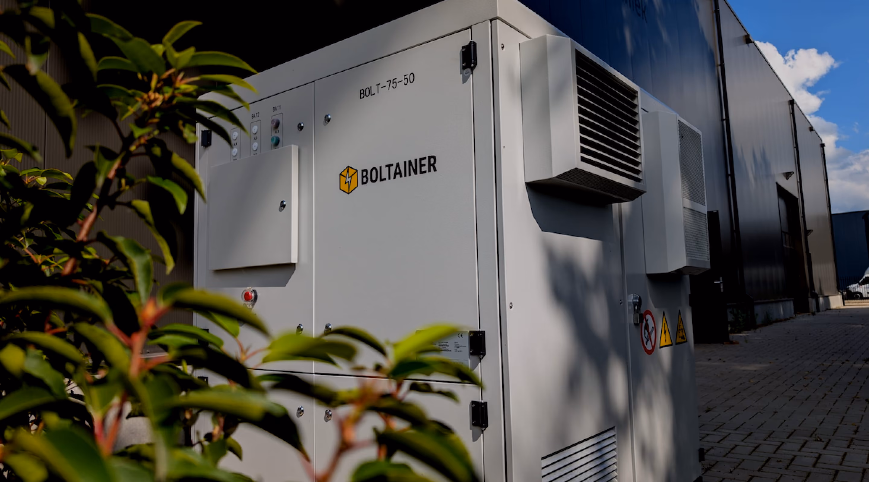 Witte Boltainer BOLT-75-50 batterijopslagsysteem met ventilatieroosters, opgesteld langs een bedrijfsgebouw onder een blauwe lucht.