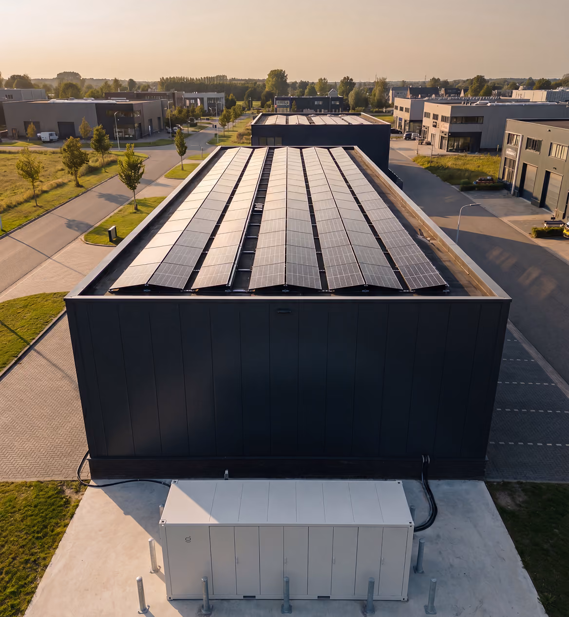 Luchtfoto van een bedrijfspand met een vol zonnepanelendak en een witte batterijopslagcontainer ernaast op een industrieterrein.