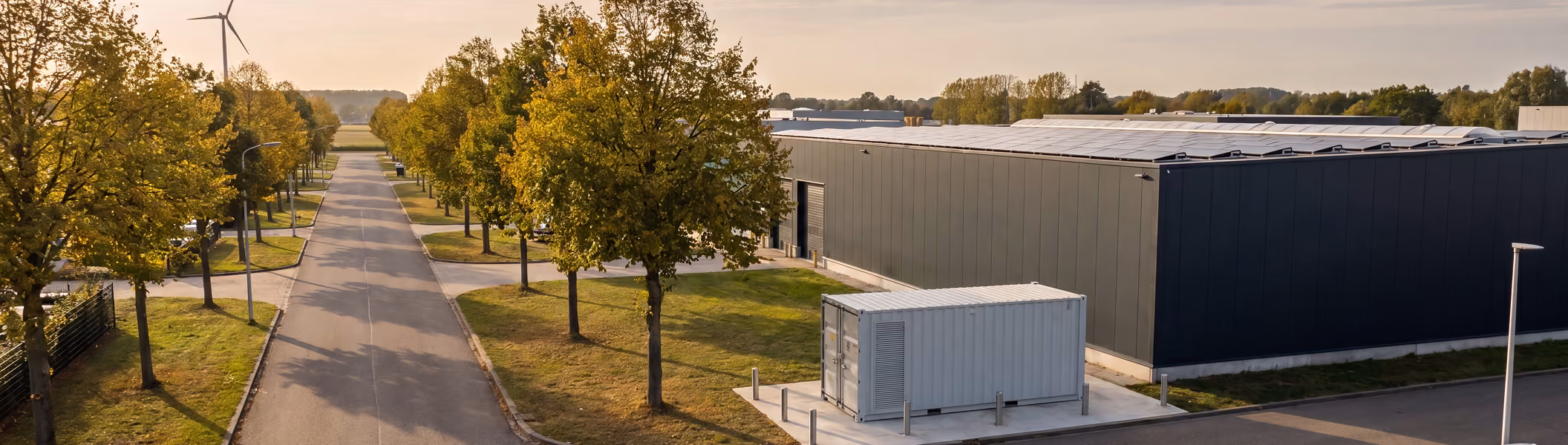 Grijze batterijcontainer op een industrieterrein naast een groot bedrijfspand met zonnepanelen, met bomen en een windturbine in de omgeving.