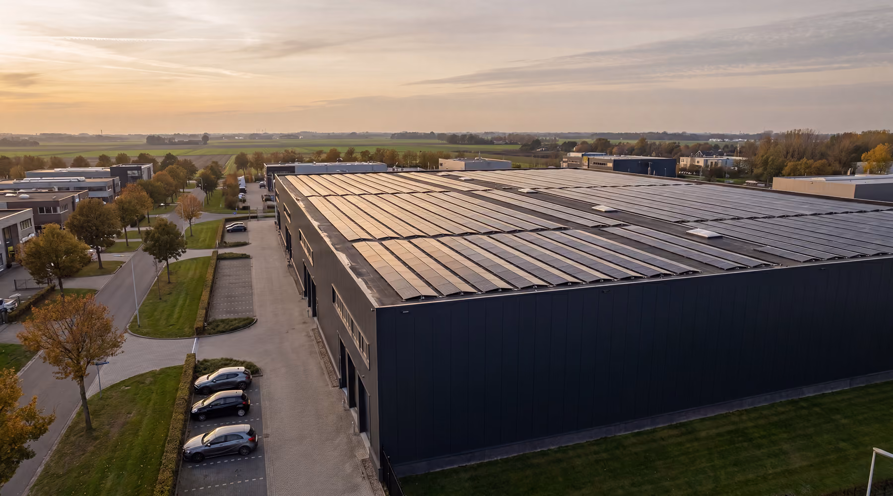 Luchtfoto van een groot bedrijfspand met antracietkleurige gevel en een dak vol zonnepanelen, met geparkeerde auto's, een bedrijventerrein en akkers in de omgeving bij zonsondergang.