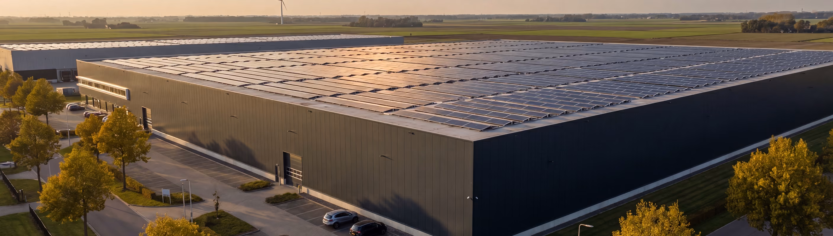 Luchtfoto van een groot industrieel pand met een uitgestrekt dak vol zonnepanelen, omgeven door akkers en een windturbine op de achtergrond in avondlicht.