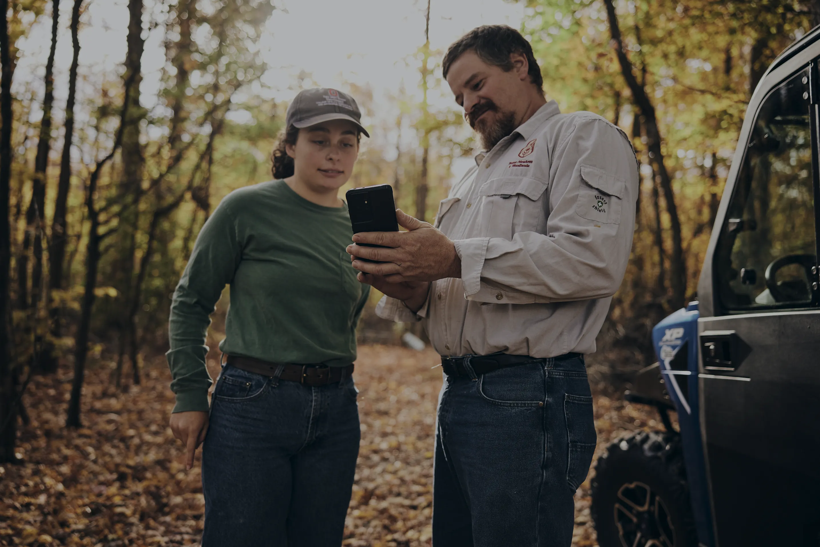 Two people standing on a forest path covered with fallen leaves, looking at a smartphone held by the man.