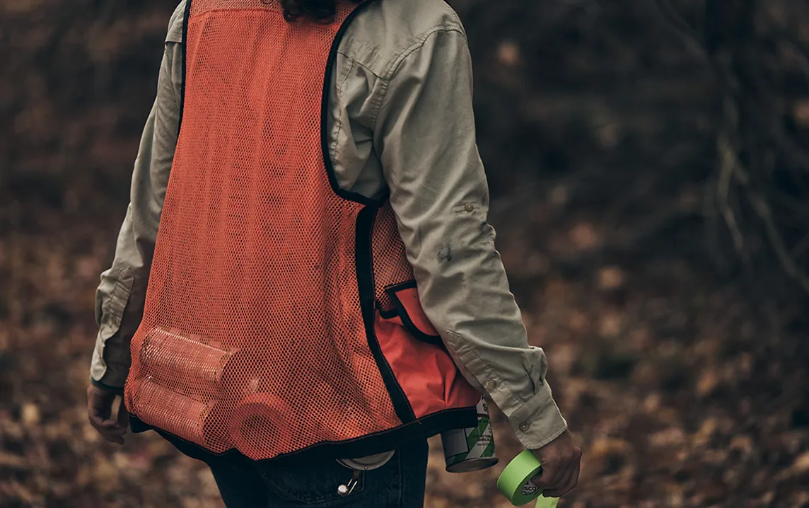 Person wearing an orange mesh safety vest holding green tape rolls and a can in an outdoor wooded area.