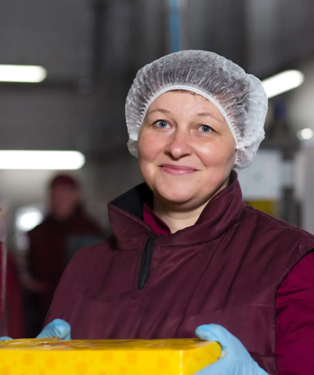 A smiling worker lady in a dairy facility wearing a burgundy uniform, blue gloves, and a hairnet, holding a bar of cheese in yellow packaging.