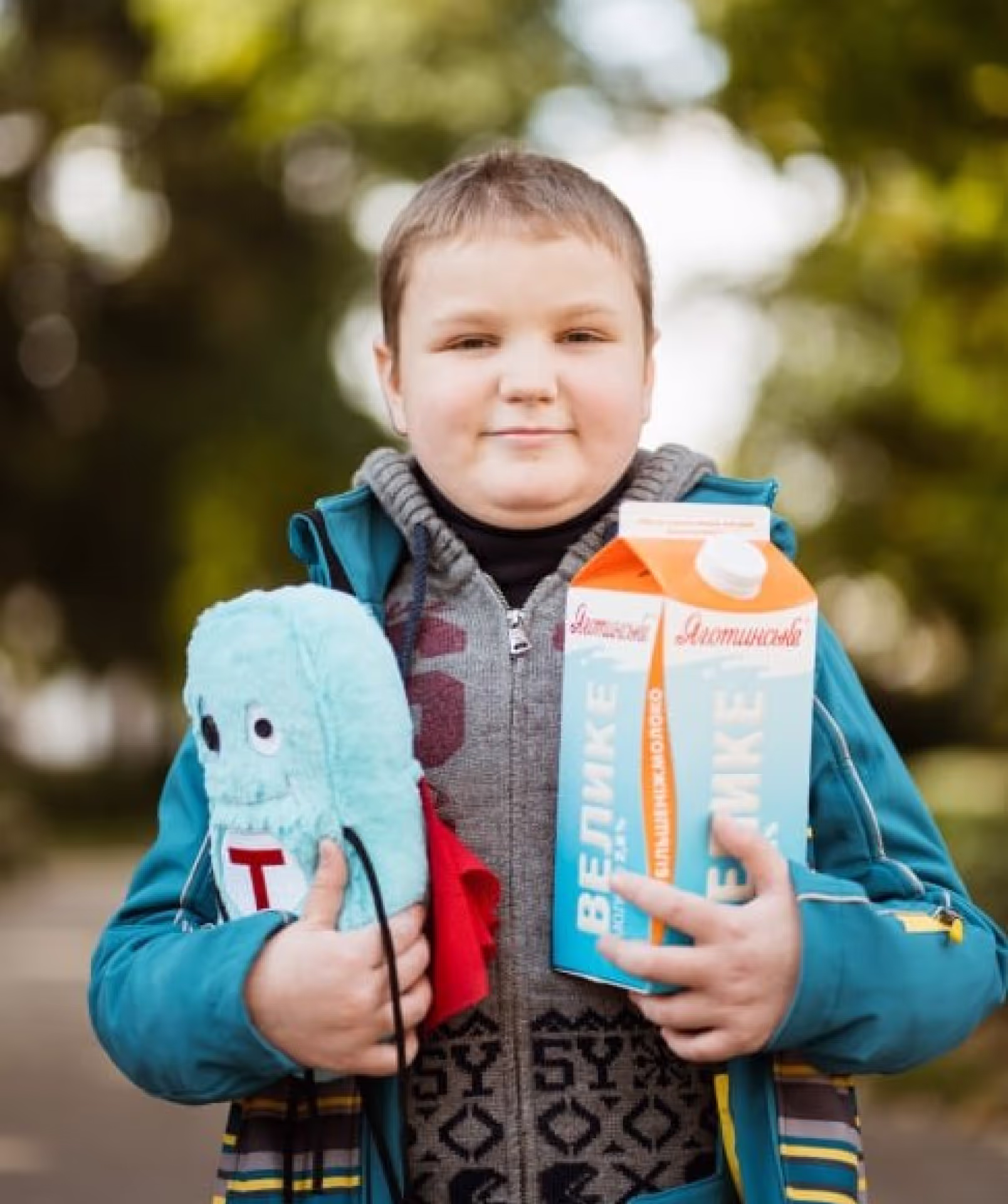 A smiling boy outdoors holding a large carton of ‘Яготинське ВЕЛИКЕ’ (Yahotynske VELYKE) milk in one hand and a plush toy in the other. He is dressed in a warm jacket and scarf, standing against a blurred background of trees.