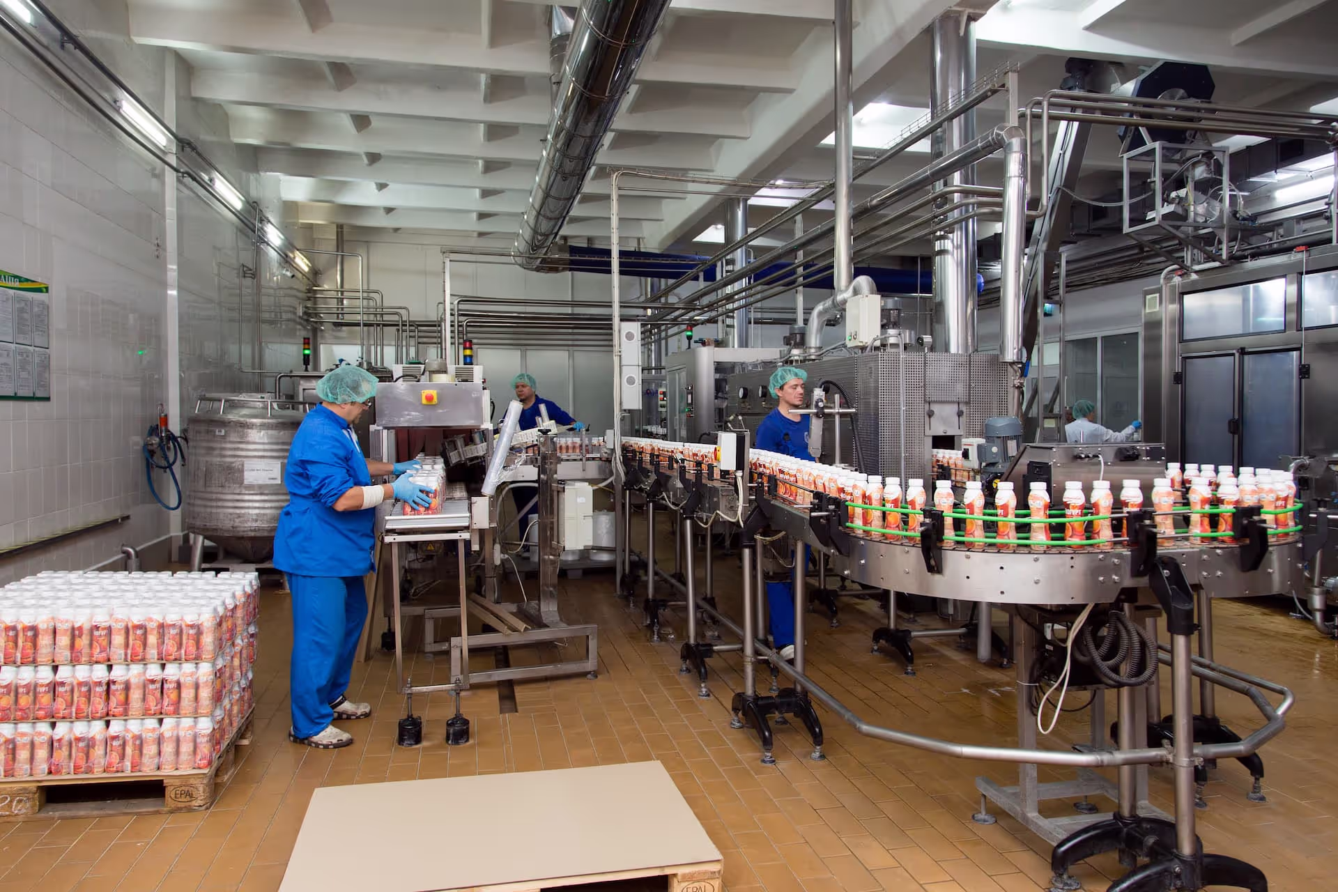 Workers in blue uniforms, hairnets, and gloves operating a dairy bottling production line. Plastic bottles of flavored dairy drinks are moving along a conveyor system, being inspected and packed in a factory equipped with stainless steel pipes and automated machinery.
