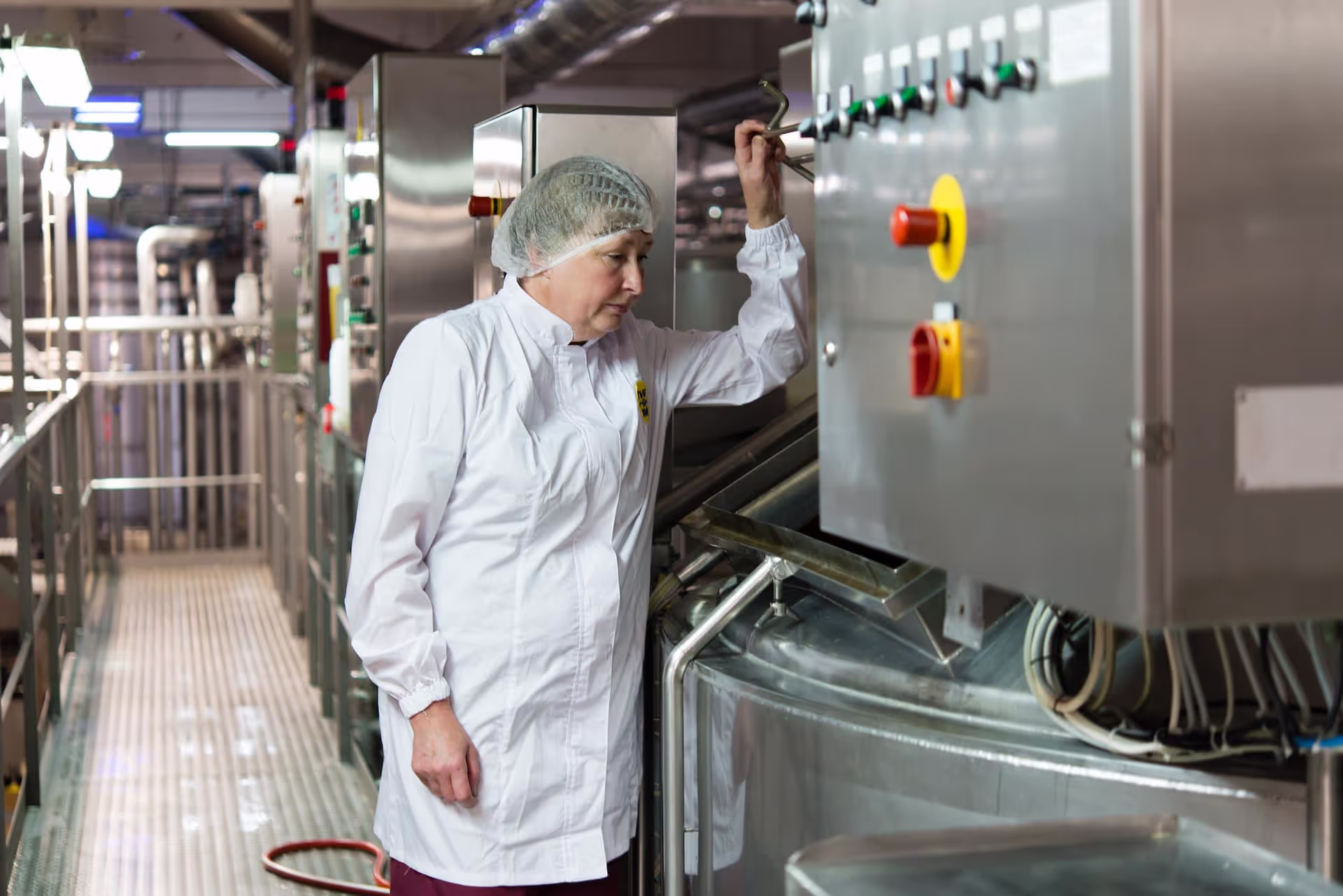 A worker in a white uniform and hairnet operating a control panel in a dairy processing facility. The stainless steel panel is equipped with various buttons, switches, and emergency stop controls, while large industrial equipment and piping are visible in the background.